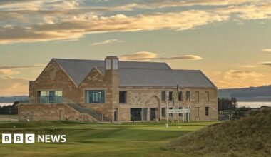 The sun has just gone down and clouds are lit in orange and gold colours above the new clubhouse at Royal Dornoch. It is a large building made of sandstone with a slate roof. A golf tee point is located in front of the building.