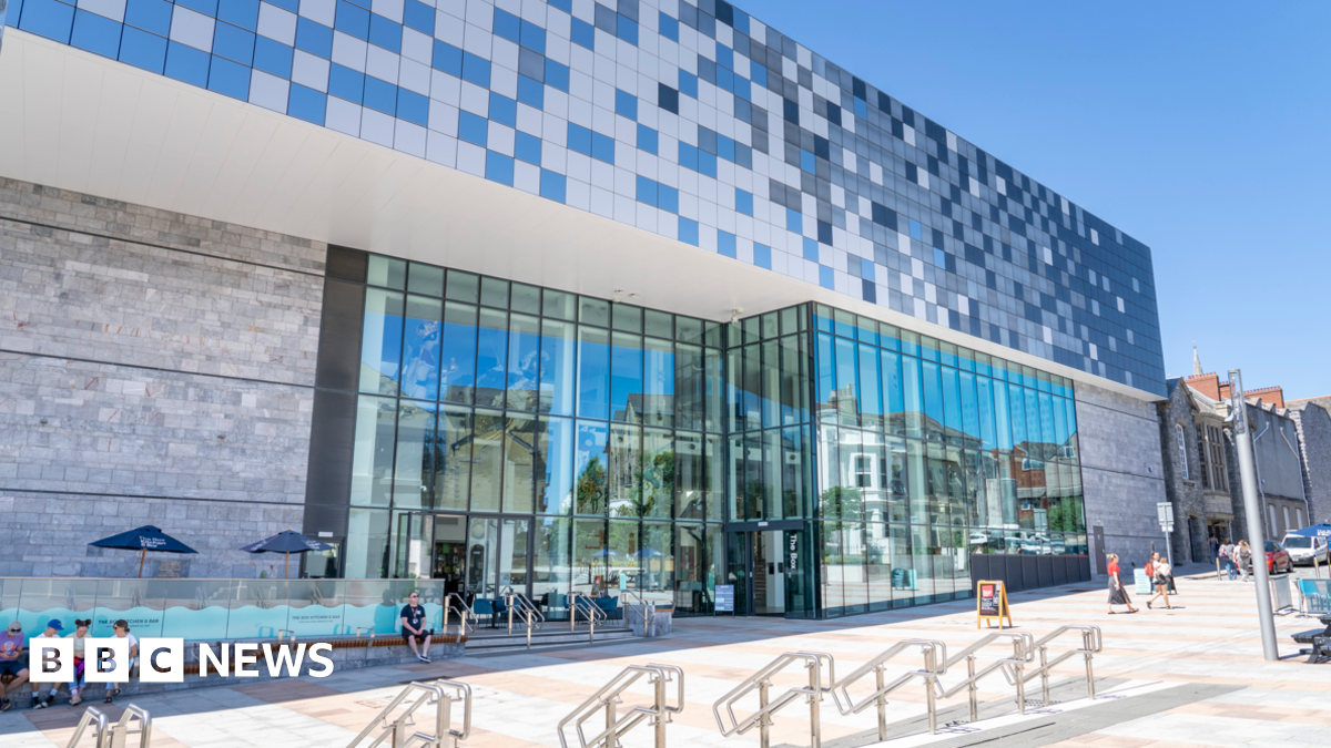 The exterior of The Box museum and art gallery. The ground floor has large glass panel windows. There are people sat outside of the museum and people walking to the right. There is a blue square design above the windows.