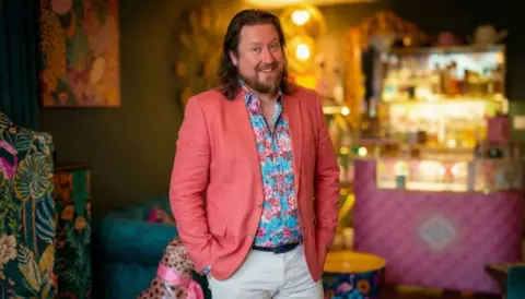 Chris Beaumont A man, Chris Beaumont, wearing a salmon pink coloured blazer and blue and red flowery shirt, poses in his home. His home is similarly colourful featuring a mixture of patterns on furniture including a sofa and bar area 