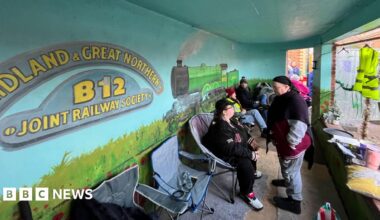 A group of people sit in portable chairs inside a bus shelter with a mural of a steam train on the wall