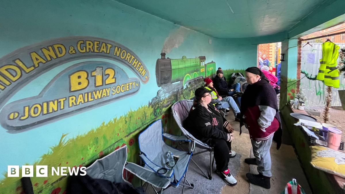 A group of people sit in portable chairs inside a bus shelter with a mural of a steam train on the wall