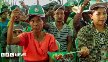 A child sitting on the ground in the aisle between two rows of chairs at a campaign rally in Mandalay. He is looking away from the stage, towards the camera.