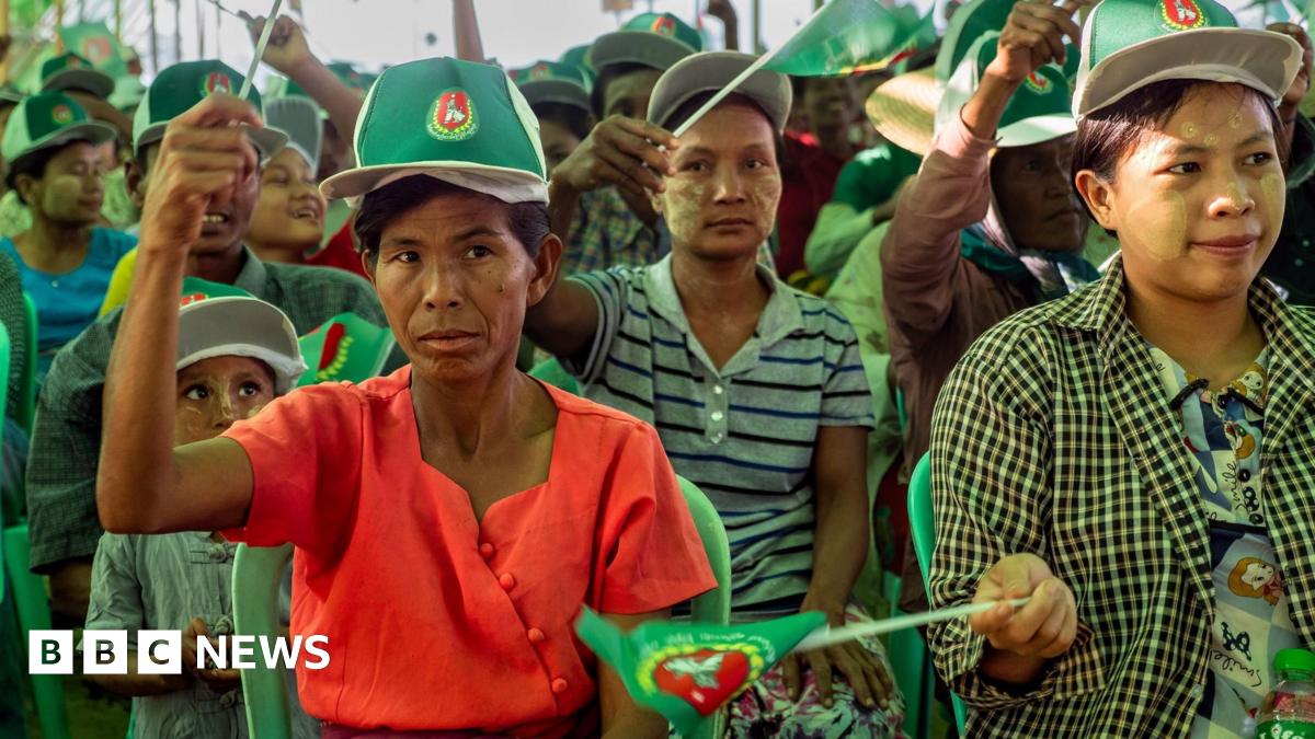 A child sitting on the ground in the aisle between two rows of chairs at a campaign rally in Mandalay. He is looking away from the stage, towards the camera.