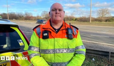 Traffic officer Robert Oates is standing by the M1. There are cars behind him on the motorway. He is wearing a fluorescent jacket with a radio and a bodycam attached to it.