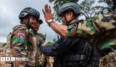 Two troops in Cambodian military uniform and helmets face off with a troop in black uniform and helmet, while in the foreground another soldier in military uniform holds out his hand with the palm acing outwards