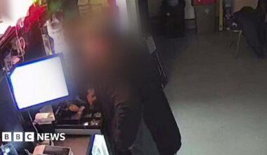 A man, whose image is blurred so he is not identifiable, is seen putting his hand into an open till.