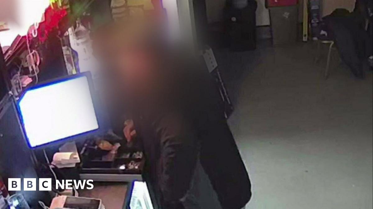 A man, whose image is blurred so he is not identifiable, is seen putting his hand into an open till.