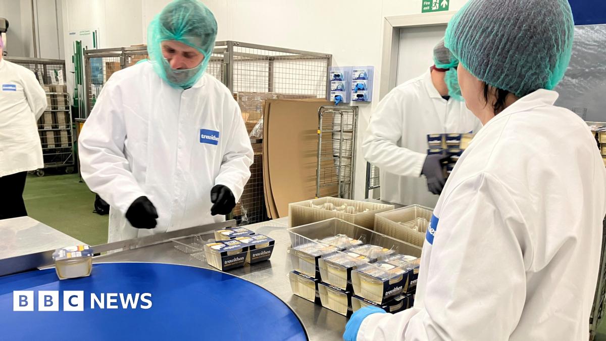 Together a man and a woman, wearing green hair nets and white overalls, are picking up tubs of clotted cream off the conveyor belt to be placed in trays at the end of the line