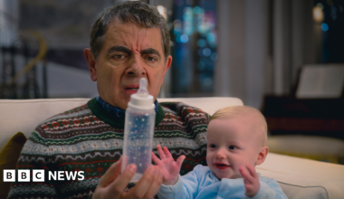 Rowan Atkinson is wearing a Christmas jumper while sitting down on a cream sofa. He is holding a baby bottle, looking confused, and has a small baby on his lap looking at the bottle.