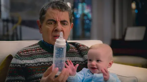 Ana Blumenkron/Netflix Rowan Atkinson is wearing a Christmas jumper while sitting down on a cream sofa. He is holding a baby bottle, looking confused, and has a small baby on his lap looking at the bottle.