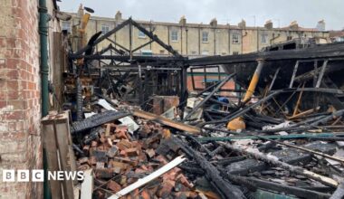 One of Underfall Yard's buildings is shown completely gutted by the fire. Just the frame of the building remains, with the roof and even exterior walls completely caved in. Piles of rubble fill the foreground.