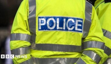 The back of a person wearing a high-visibility jacket, which is bright yellow with reflective silver stripes across the arms and torso. On the back of the jacket, there is a large rectangular patch with the word "POLICE" written in bold, white capital letters on a blue background. The reflective stripes are positioned horizontally.