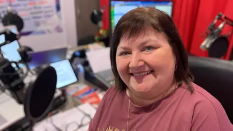 BBC A woman with chin length brown hair is smiling at the camera. She is wearing a plum coloured t-shirt and is sitting in front of a radio studio