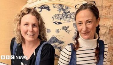 Karen Evans, with blonde curly hair, wearing a black top, blue tights and black boots, holding a cushion with the artwork on. The cushion is white and has blue illustrations on it. Sitting next to her is Maddy Humberstone, holding a similar cushion, with brown hair tied back, wearing jeans and a white stripey top and Converse trainers.