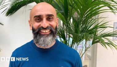 Navraj Singh is standing in front of a plant and a window at home. He is bald with a beard and is wearing a blue jumper. He is smiling and looking happy.