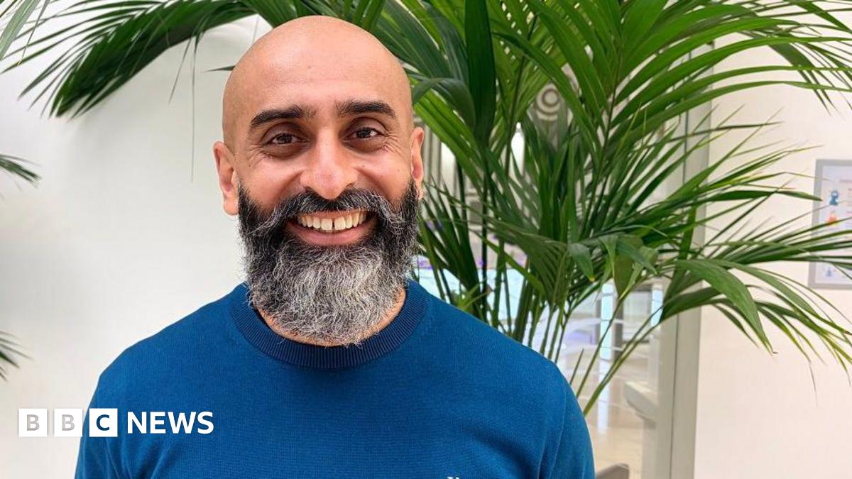 Navraj Singh is standing in front of a plant and a window at home. He is bald with a beard and is wearing a blue jumper. He is smiling and looking happy.