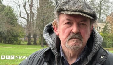 An older man wearing a dark jacket with a fleece-lined hood and a flat cap is standing outdoors.