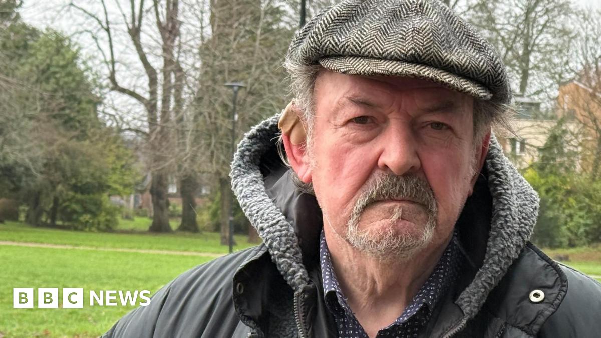 An older man wearing a dark jacket with a fleece-lined hood and a flat cap is standing outdoors.