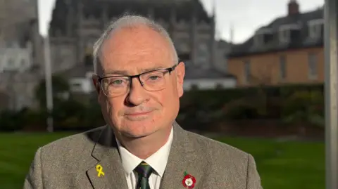 Steve Aiken's mouth is closed, his lips gently drawn in. He is wearing square-shaped glasses, black-rimmed at the top and without a rim at the bottom. He is wearing a tweed jacket, with has a pin on either lapel. behind him is an old stone building, perhaps a church. 