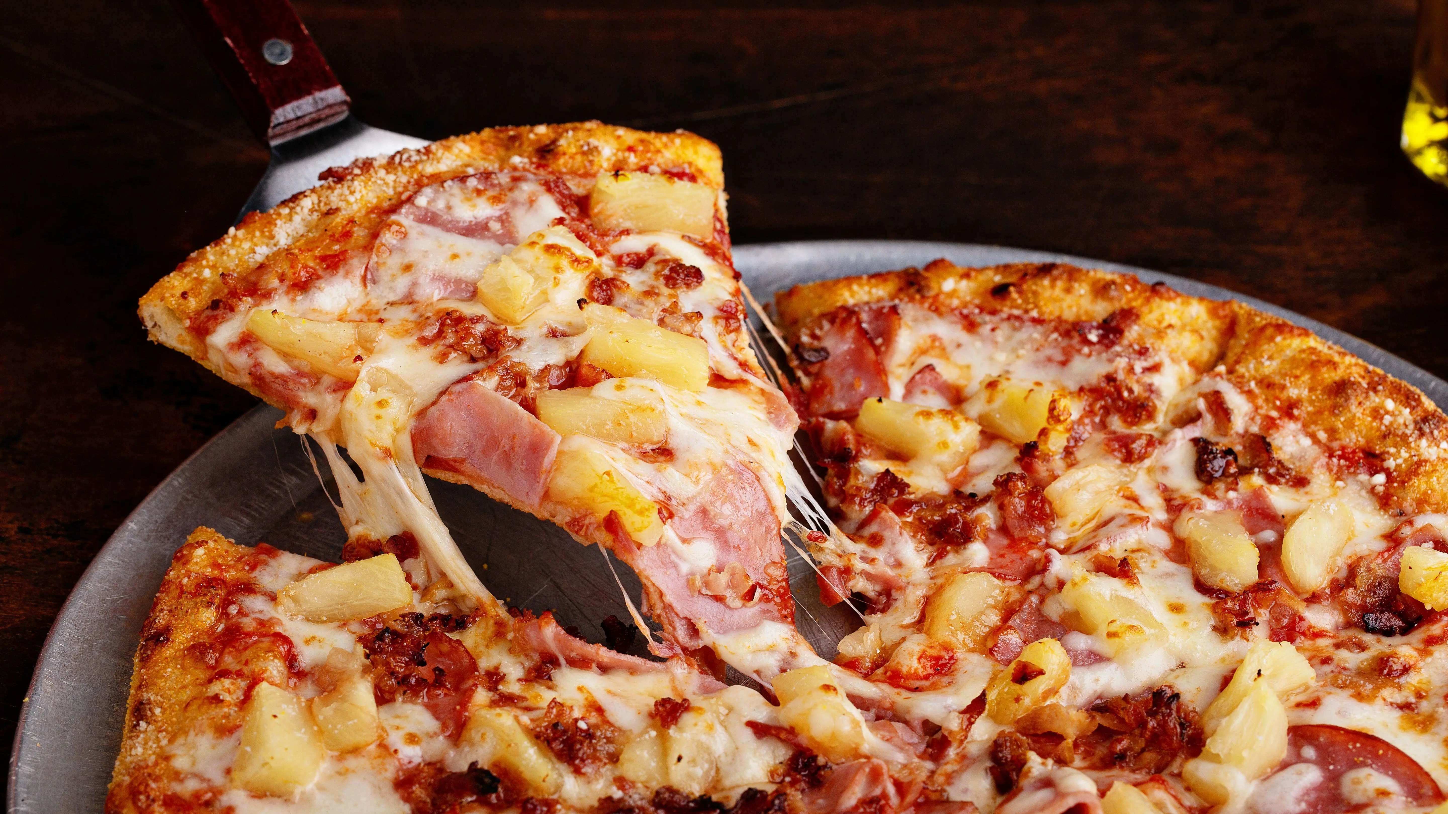 Hawaiian pizza with ham and pineapple, with a slice being lifted and cheese stretching.
