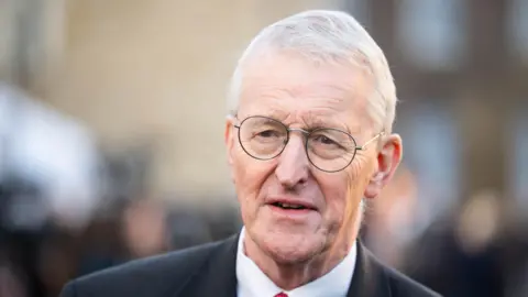 PA Media Secretary of State Hilary Benn is wearing round glasses with thin rims, and his thinning grey hair is swept to the side.
