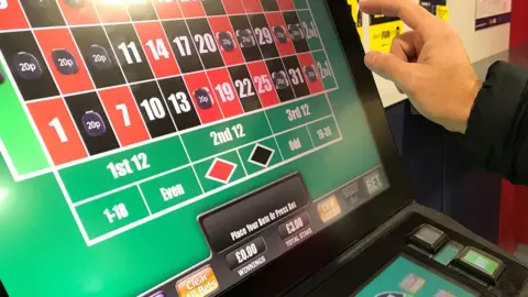 Reuters A man's hand is pictured about to press the brightly lit screen of a slot machine. The screen shows a series of red and black squares, some of which are filled with the price of bets placed. Details of stakes and winnings are also pictured. 