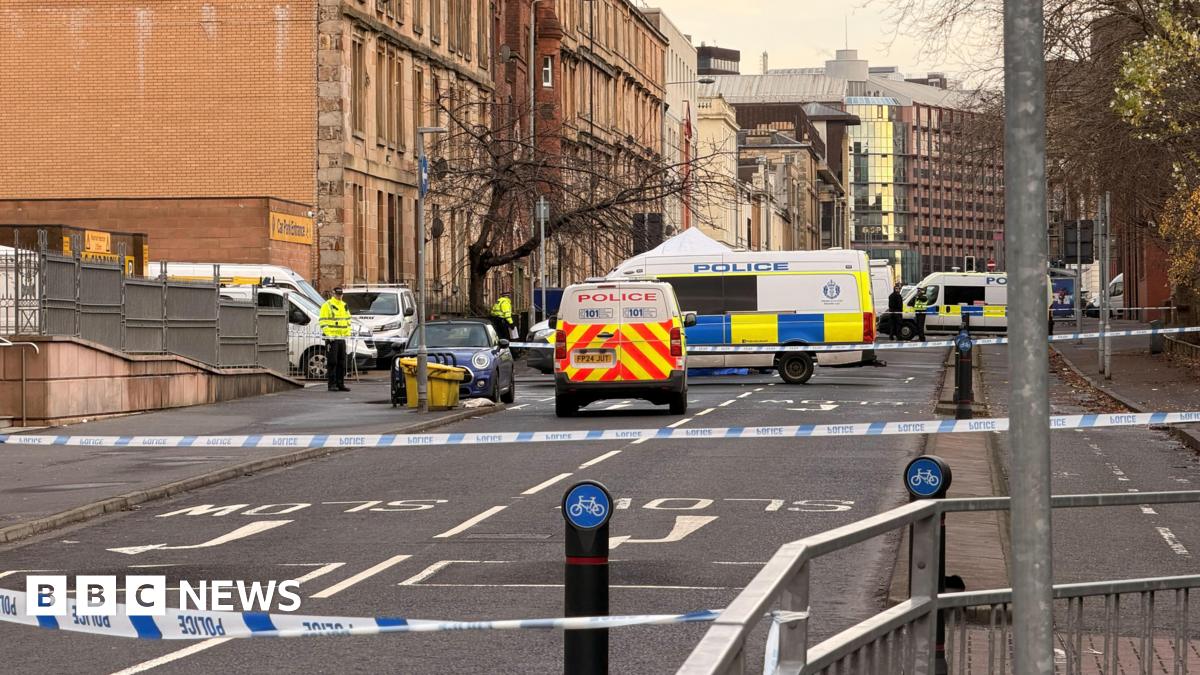 A street with a police cordon, police tape and various vans and cars parked there
