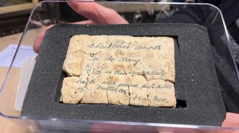 Leeds Museums A beige-coloured biscuit cracked into several pieces but arranged together to form its original rectangular shape. Faded blue handwriting is visible across the surface with an inscription that reads: 'Christmas dinner in the army. Give us this day our daily bread and please put a bit of butter on it. From Max.'
