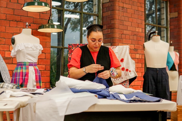 Yasmin Proctor-Kent and sewing something on the set of The Great British Sewing Bee, surrounded by mannequins, fabric and a waistcoat with a poppy design on the board behind her