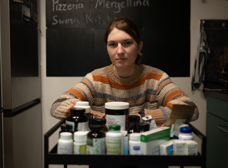 A woman in a striped knitted sweater looks at the camera from behind a cart loaded with medication bottles in her apartment.