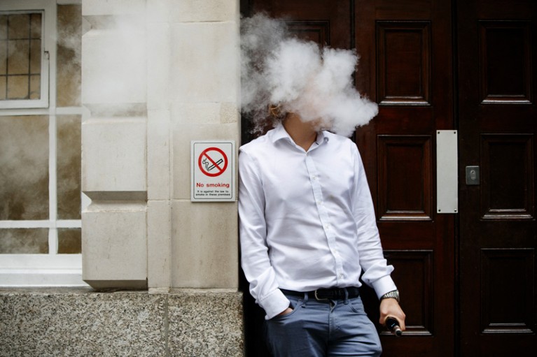 A man in a shirt standing in a doorway next to a no smoking sign, exhaling a large cloud of vapour.