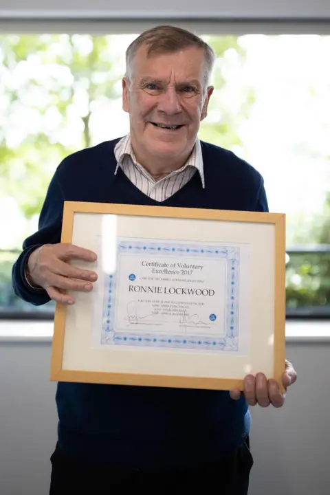Rob Parsons Ronnie is pictured holding a certificate for his work volunteering. The certificate is a large wood frame which Ronnie holds infront of my chest. He wears a navy v neck jumper, white shirt and smiles at the camera. Behind him is a window which is full of blurred green trees. 