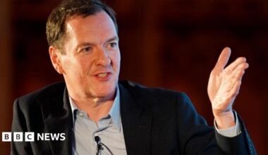 George gestures with his hand while looking positive. He's wearing a black suit jacket and a grey shirt.