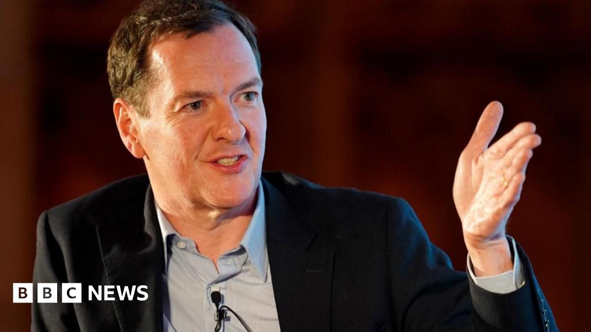 George gestures with his hand while looking positive. He's wearing a black suit jacket and a grey shirt.