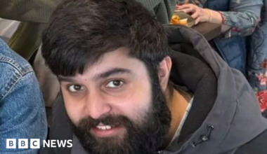Adam Ali Hussain, a south Asian man in his 20s with black hair and a medium length beard. He is smiling and looking up towards the frame. He is wearing a black and grey winter coat