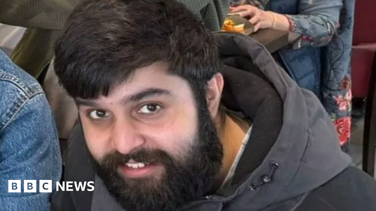 Adam Ali Hussain, a south Asian man in his 20s with black hair and a medium length beard. He is smiling and looking up towards the frame. He is wearing a black and grey winter coat