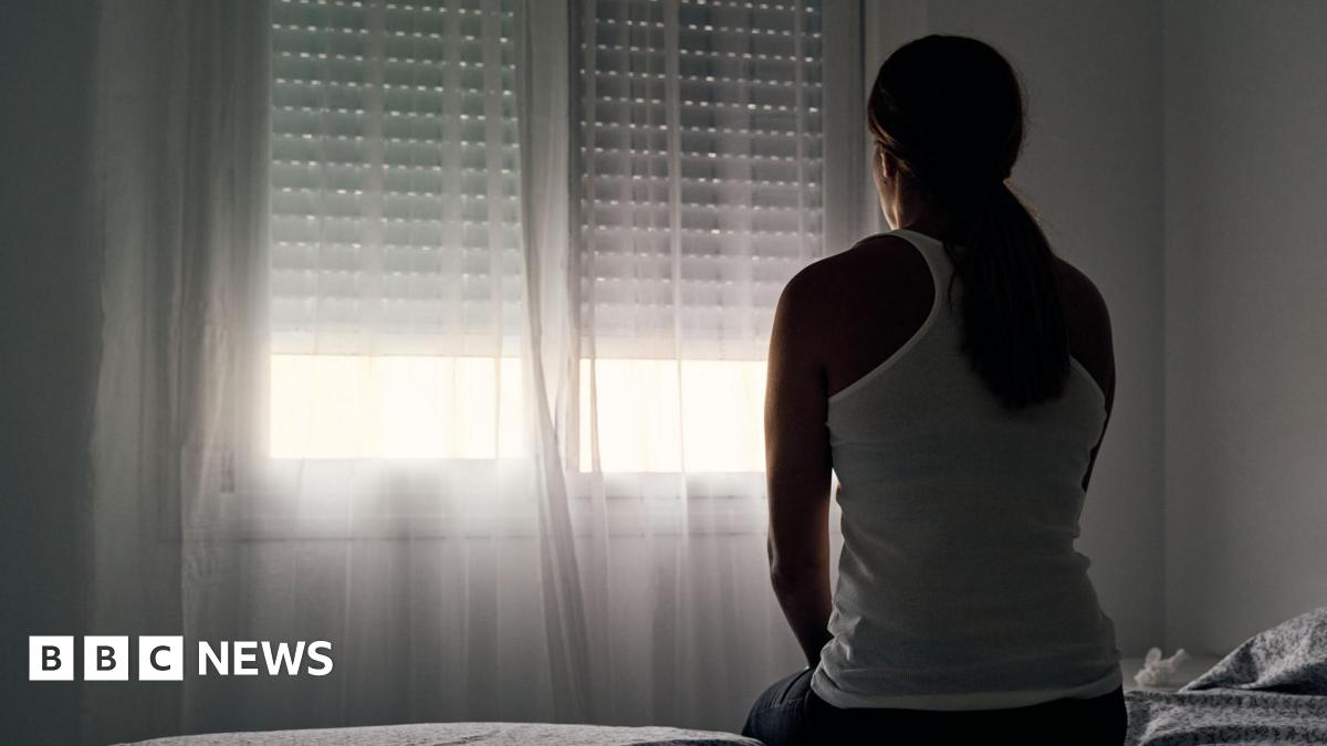 A woman wearing a white vest top with her hair tied back sits with her back to the camera, looking out the window.