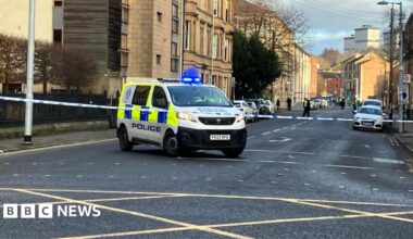 A city street, with police tape covering part of the area. A police van is parked at the tape, and officers can be seen in the distance.