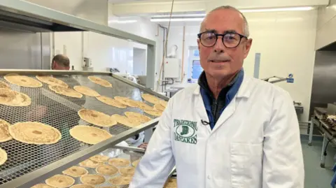 A man in glasses is wearing a white overcoat with the words Traditional Oatcakes on the front. He is next to a large rack of the food