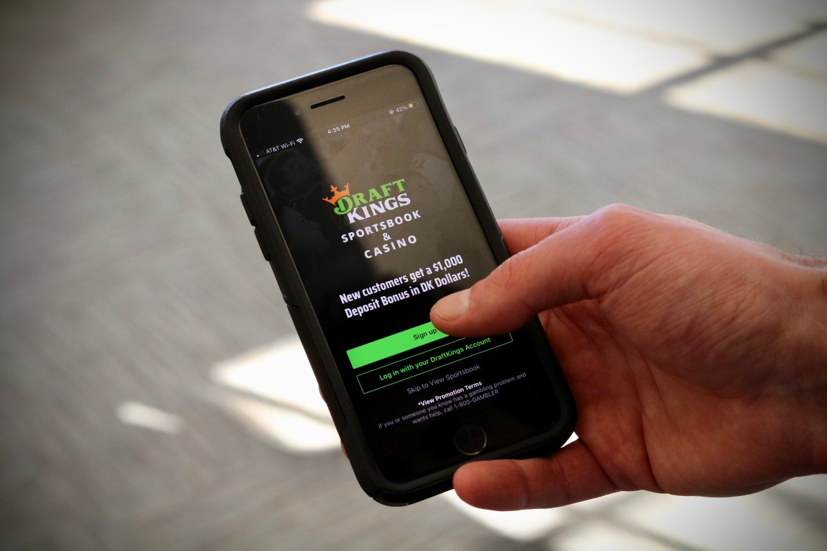 A person holding a smartphone showing the DraftKings Sportsbook & Casino app's login screen, which offers a $1,000 deposit bonus for new customers.