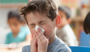 Child sitting indoors with hands near mouth, wearing a light blue top, in a classroom setting with other children and colorful background elements.