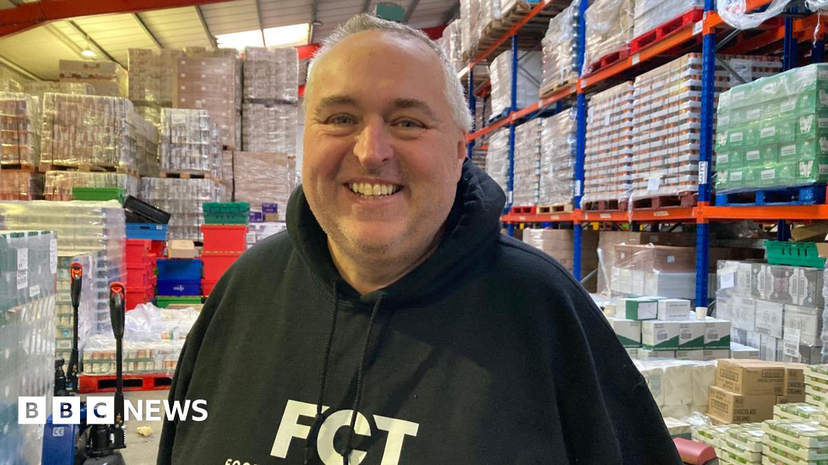A man wearing a dark hoodie stands in a warehouse full of pallets containing food products.