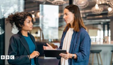 Two young business women working together, standing at modern office space room looking at a tablet computer. Both dressed in smart-casual business clothing.