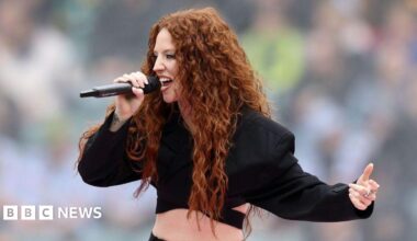 Jess Glynne is singing into a mic, she is wearing a cropped black suit jacket. SHe has long curly ginger hair.