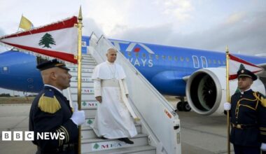 A man in white religious robes and skullcap descends the steps of an airliner as soldiers in dress uniform stand guard with flags.