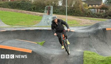 A man cycles on the pump track, towards the camera. He is wearing black clothes and a grey helmet and is riding an orange bike. The pump track has orange stripes and there is grass and houses behind it.