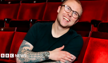 Joe in a black top, wearing glasses, sitting in red theatre/cinema seats