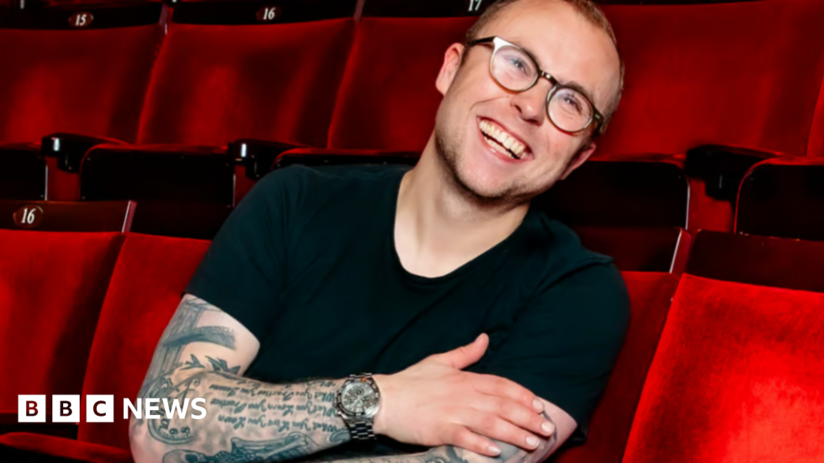 Joe in a black top, wearing glasses, sitting in red theatre/cinema seats