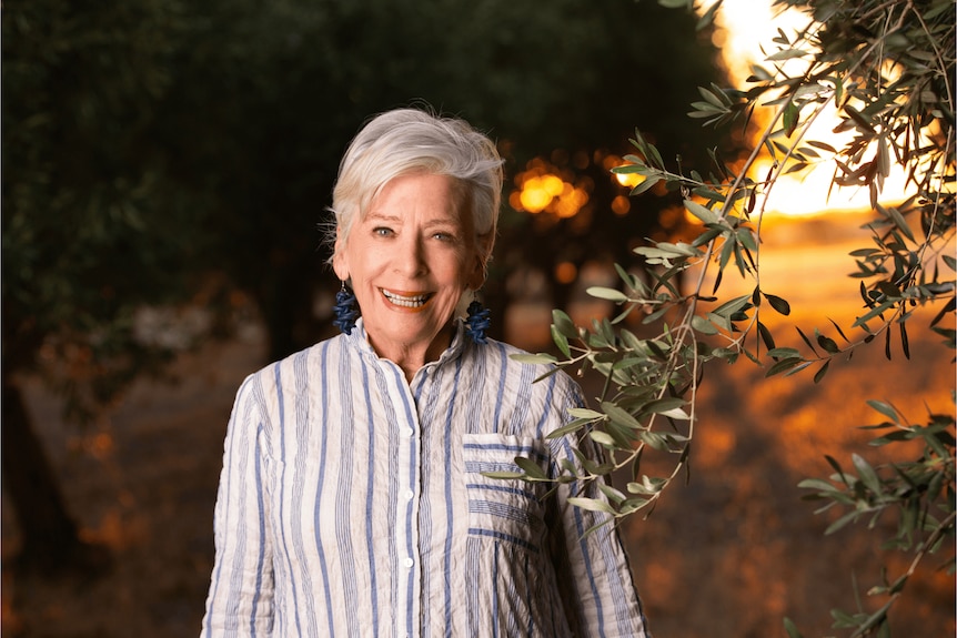 Maggie Beer smiles at the camera while standing in a grove of trees, a bright sunset lighting the field behind her. 
