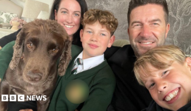 A woman and man, both with dark hair, sat on a sofa in front of a grey wallpaper backdrop. In front of them are two blonde boys, one with curly hair and the other with straight, and a brown cocker spaniel. All four people are looking at the camera and smiling.
